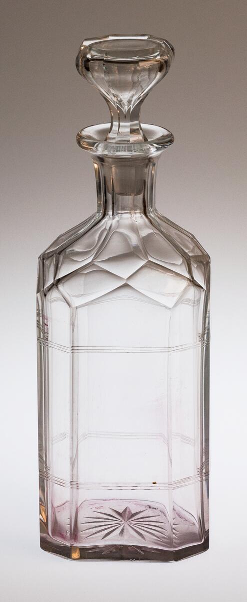 AV GLAS, MED PROPP. ÅTTKANTIG, RUNT BOTTEN MED STJÄRNSLIPNING. 2 GGR 3 RÄNDER PÅ TVÄREN PÅ BUKEN. OREGELBUNDEN FACETTSLIPNING PÅ AXELPARTIET. HALSEN FACETTSLIPAD. UTVIKT MYNNINGSKANT. PROPPEN BLÅST MED BUBBLA, PLAN OVANSIDA, SLIPAD ÅTTKANTIG, H. 25, BR. 8,7 CM.