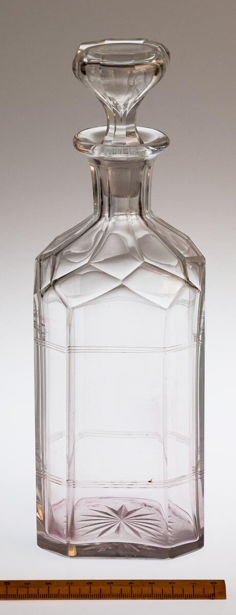 AV GLAS, MED PROPP. ÅTTKANTIG, RUNT BOTTEN MED STJÄRNSLIPNING. 2 GGR 3 RÄNDER PÅ TVÄREN PÅ BUKEN. OREGELBUNDEN FACETTSLIPNING PÅ AXELPARTIET. HALSEN FACETTSLIPAD. UTVIKT MYNNINGSKANT. PROPPEN BLÅST MED BUBBLA, PLAN OVANSIDA, SLIPAD ÅTTKANTIG, H. 25, BR. 8,7 CM.