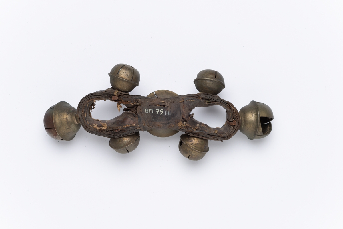 Bøyd lærrem (8-tallsformet), påsatt 7 kuleformede hule metallbjeller i forskjellige størrelser. Bjellene har spalter og innlagt kule som klirrer når den rister. For plassering oppå bogtrærne.