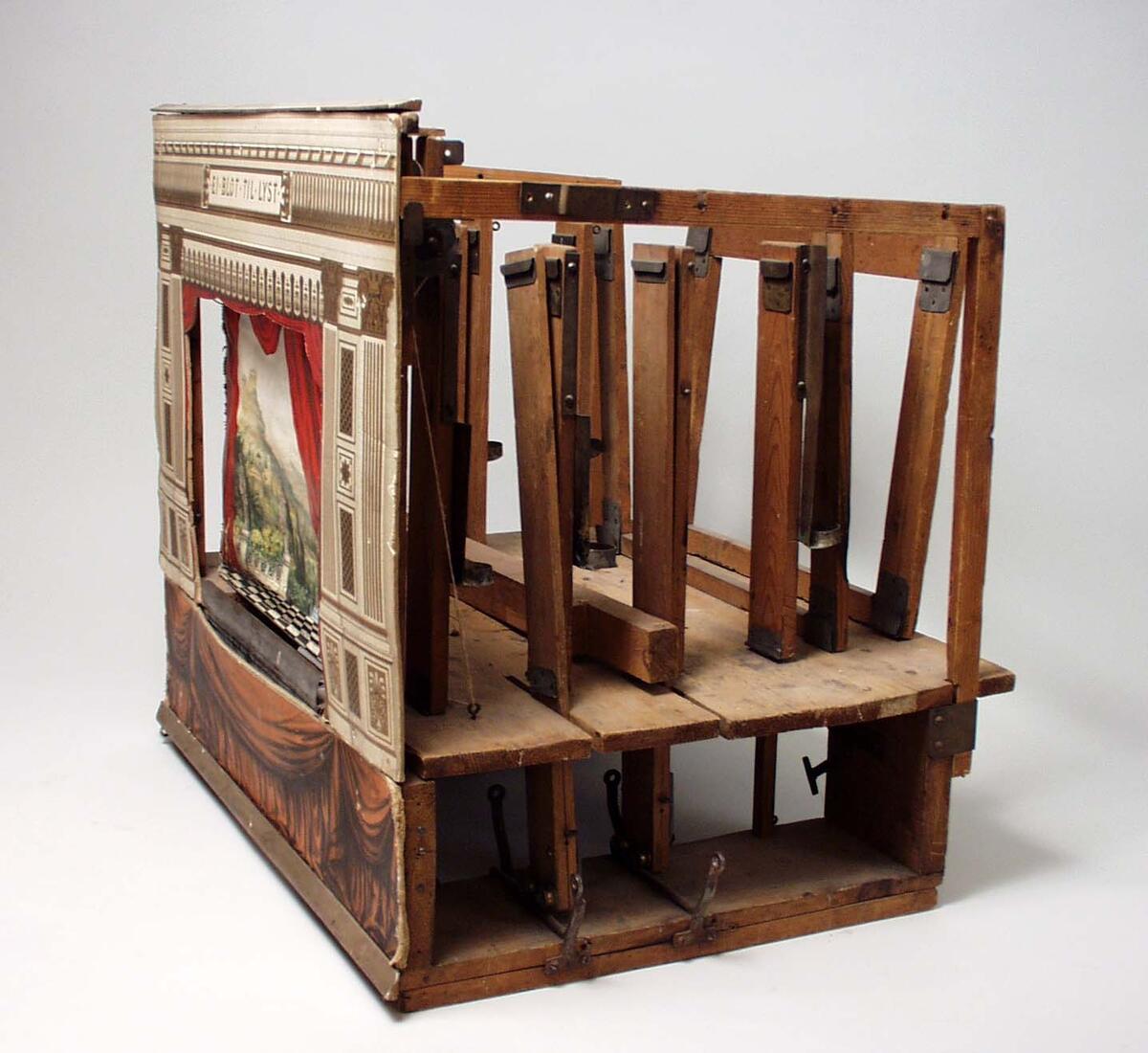 Modellteater, Alfred Jacobsen. Stomme av trä. Framsidan runt scenen klädd med flerfärgad papp, ovanför scenen texten: "EI BLOT TIL LYST". Ridå av papp föreställande Akropolis, tre änglar längs ned t v och sex t h, rödfärgade draperier, troligen Alfred Jacobsens Teaterdekorationer Nr 102. Till dockteatern hör en mängd delar, bl a kulisser, teaterdekorationer och figurer av papp, se vidare C13410:2 - 115.

Nyreg 2001-01-22 KS