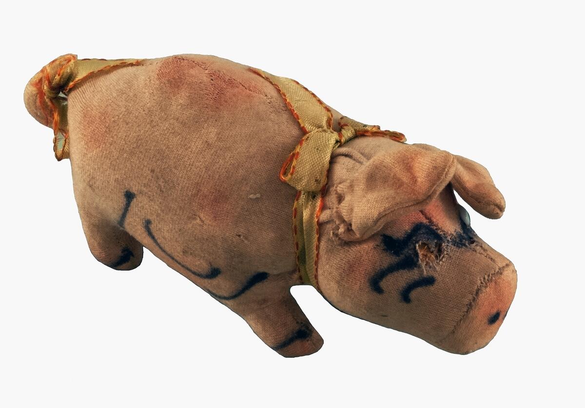 Lite leksaksgris av från början skärt tyg, ögon av glas (ett saknas), gula siden band kring hals och svans, målad dekor i svart på tryne, kropp och ben, nu ganska sliten och smutsig
nyreg data 020122 li