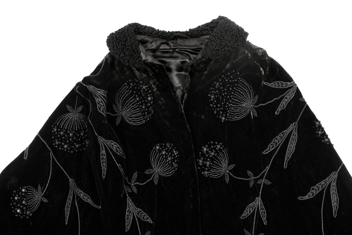 Sort kep i fløyel med lissebroderi, perler og plaststjerner (eller bakelitt?) som former blomster. Krage med persianer. Foret med silke eller kunstsilke. Den har to hemper i halsen. 