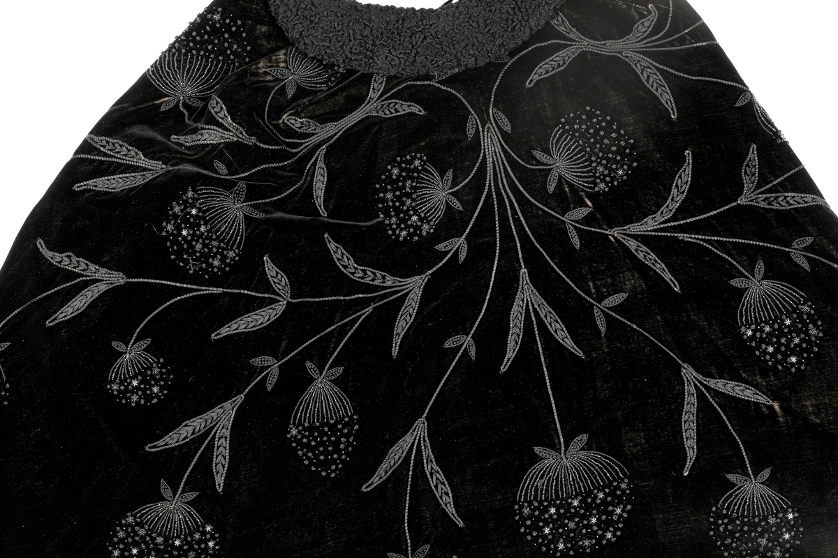 Sort kep i fløyel med lissebroderi, perler og plaststjerner (eller bakelitt?) som former blomster. Krage med persianer. Foret med silke eller kunstsilke. Den har to hemper i halsen. 