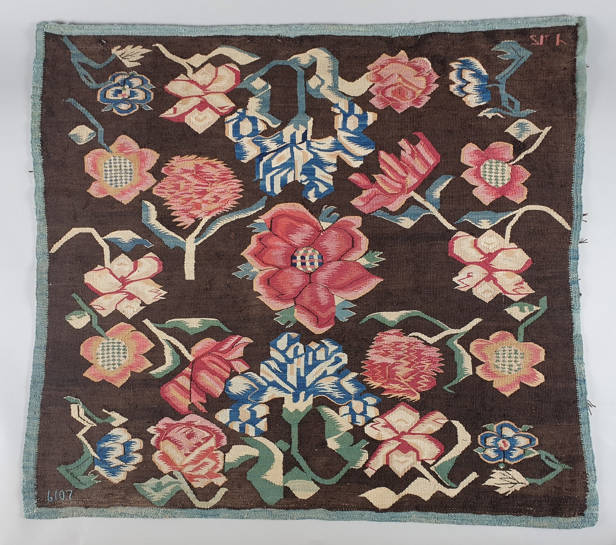 Putetrekk av billedvev. Renning av lin og innslag av ull (bomull?). Røde roser, rosa markblomster og blå liljer med grønne og hvite blader på mørk bunn.