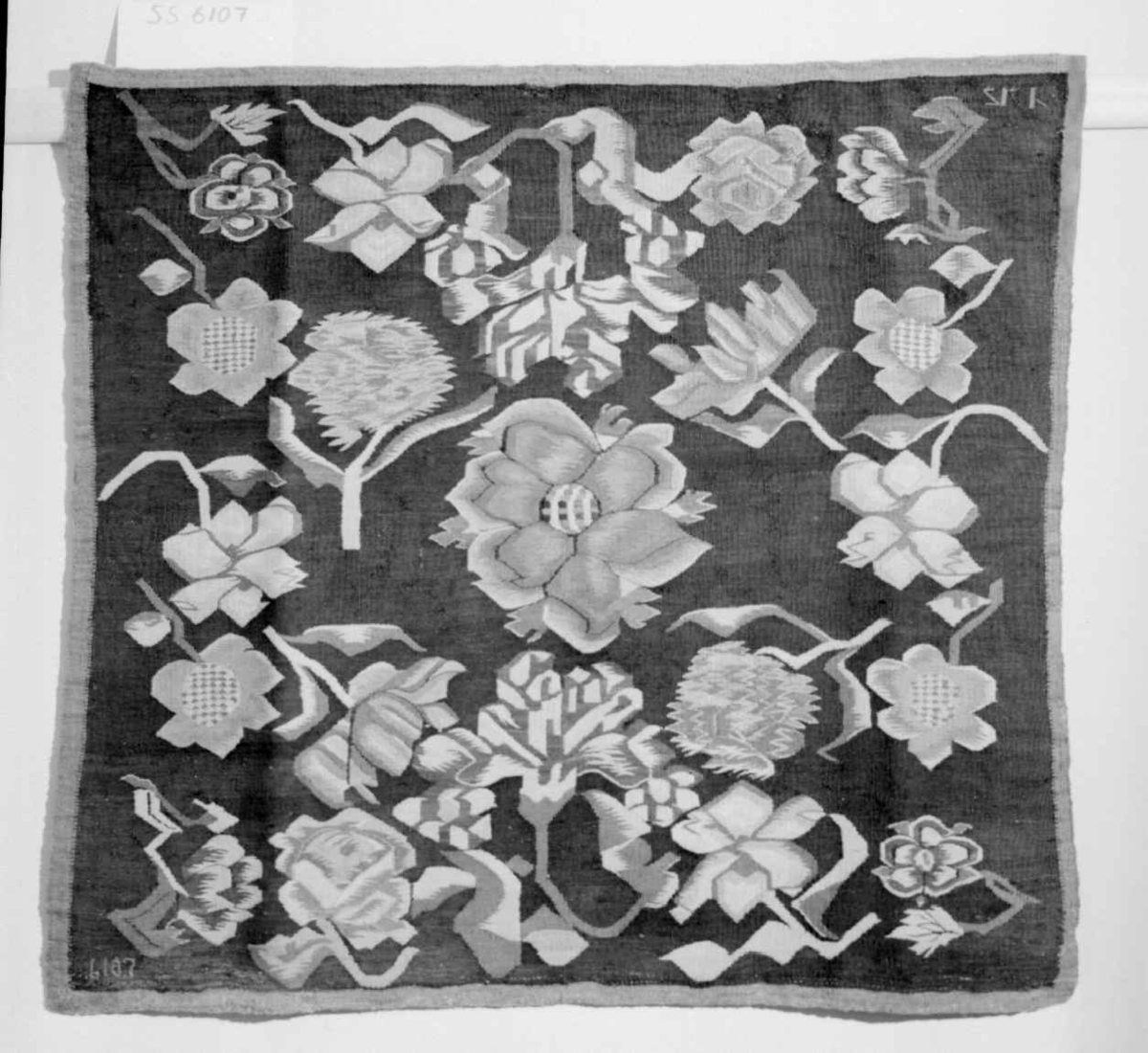 Putetrekk av billedvev. Renning av lin og innslag av ull (bomull?). Røde roser, rosa markblomster og blå liljer med grønne og hvite blader på mørk bunn.