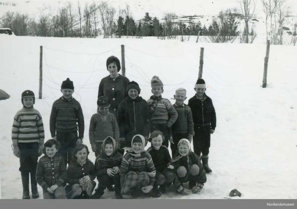 Lærer og elever ved Saursfjord skole, 1. og 2. klasse. Lærerens navn er Johanne Aasjord. Guttene fra venstre: Torbjørn Johansen, Rolf Alpøy, Kjell Gunnar Kristensen, Roald Pettersen, Henry Stensland, Herulf Abrahamsen og Valter Pedersen.  Jentene fra venstre: Lina Sofie Normann, Sylvia Pettersen, Wenche Henriksen, Gerd Pettersen, Dagny Markussen og Gerd Alpøy.