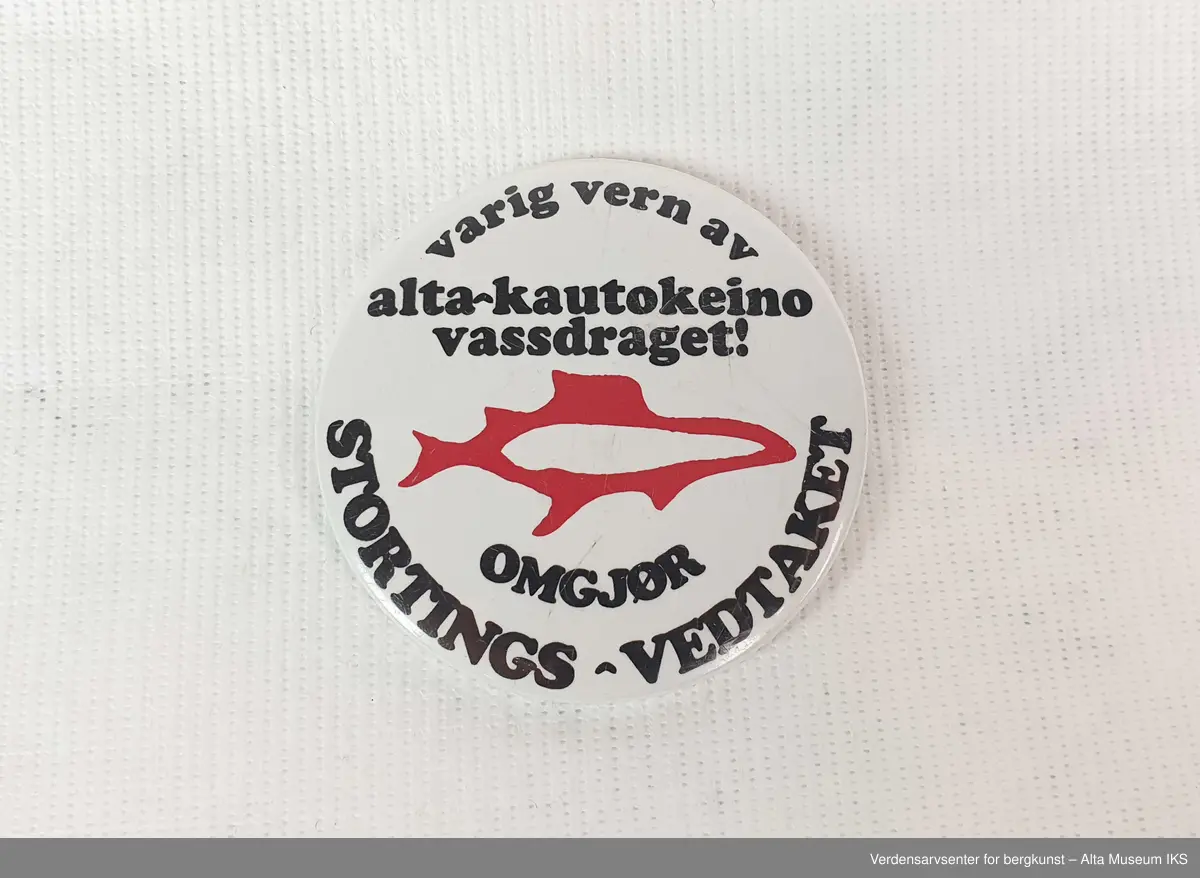 Rund hvit button i plast og metall, motivet er av en rød laks med teksten: "Varig vern av Alta-Kautokeinovassdraget! Omgjør Stortings-vedtaket".