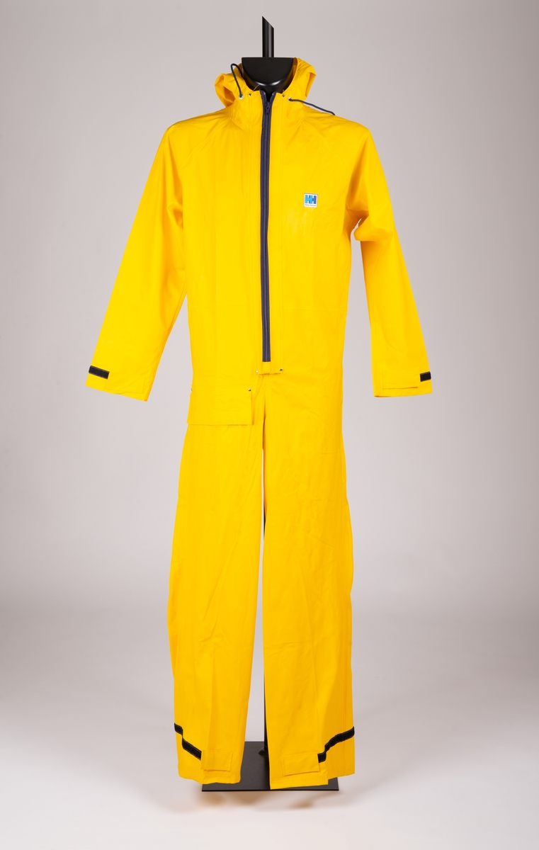Art. nr. D-651. Gul seilerdress i plastbelagt nylon. Innkjøpt metervare. Glidelås med vifte i front. En vanntett lomme med klaff og borrelås på høyre side foran. Borrelåsstramming nederst på ben og ermer. Fast hette med snortrekk. Løpegang med strikk i livet bak. Etikett i nakke.