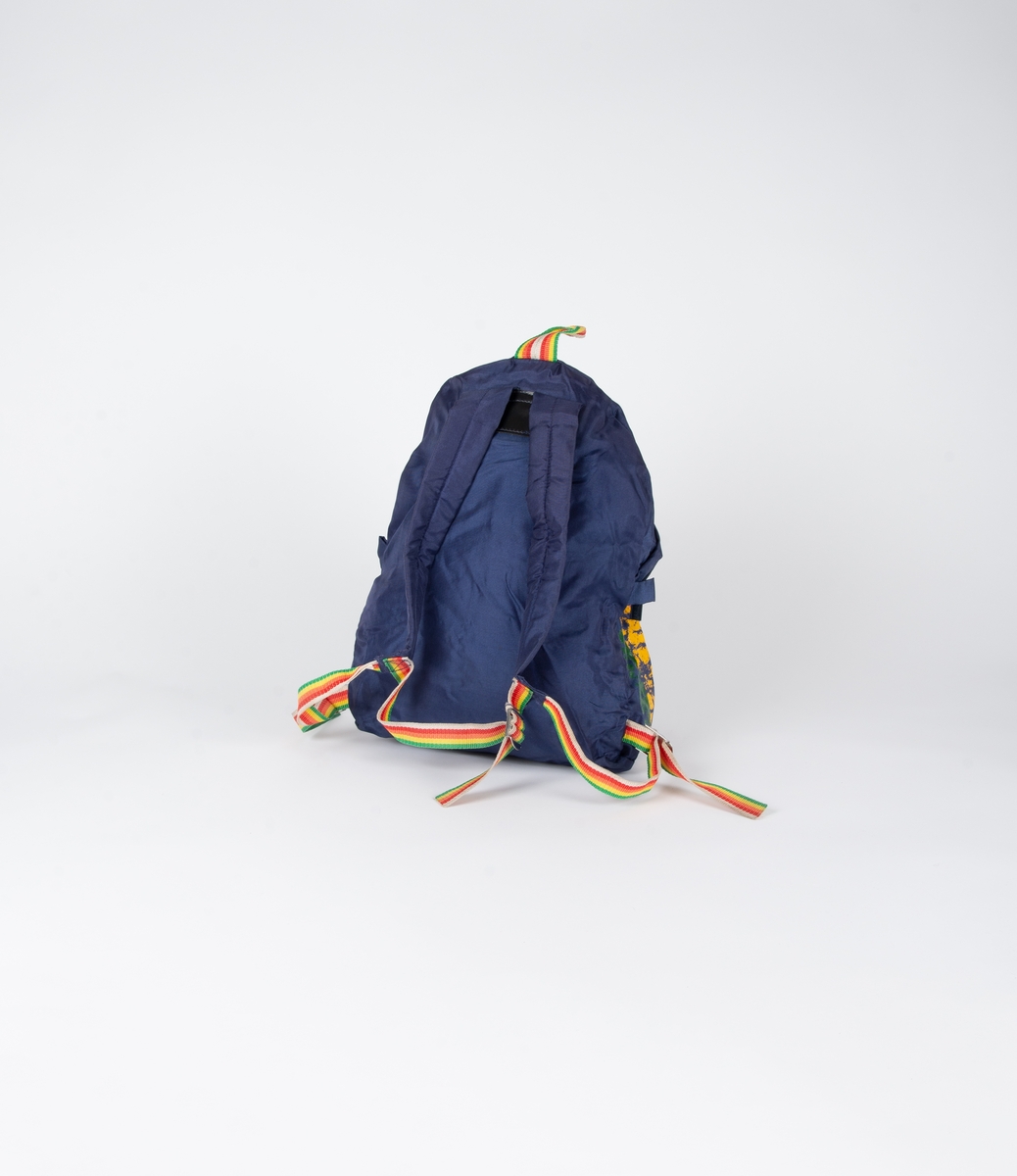 Blå ryggsekk med frontlomme med glidelåslukning. Vertikale striper i grønt, gult, oransje og hvit til høyre. Vevde bånd i regnbuens farger på seler, opphengsstropp og magebelte. Regulerbare spenner i metall. Selene har et polstret stykke øverst. Selene er festet med et svart skai-stykke med to niter øverst på ryggen. Vevde hemper, en på hver side, antagelig til karabinkroker.