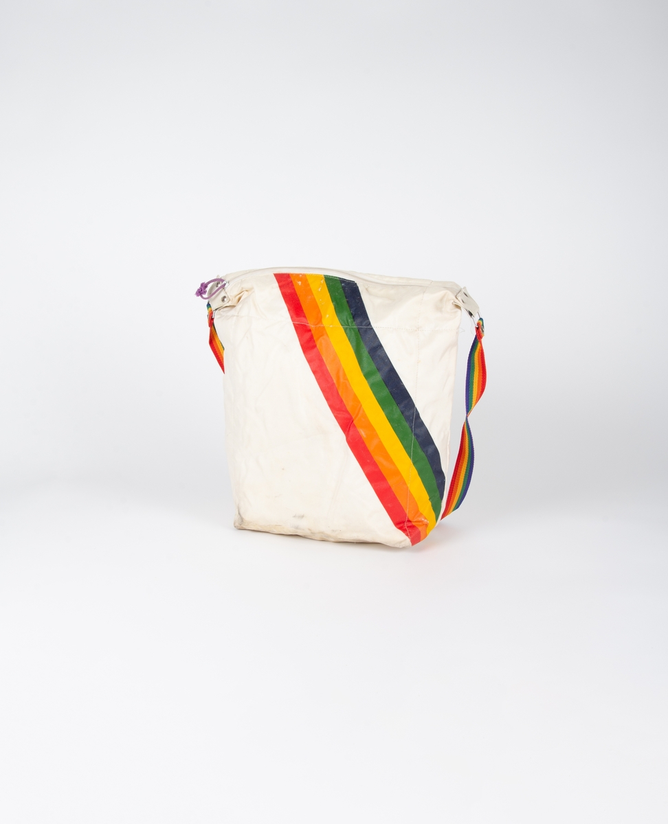 Hvit bag med skråstilte regnbuestriper. På den andre siden er det ni repiterende logoer nedover: Helly-Hansen. Glidelåslukning på toppen. Bærerem vevd i regnbuestripe på hver side festet med tre niter på forsterkningsstykke. Metallring mellom rem og forsterkningsstykke.