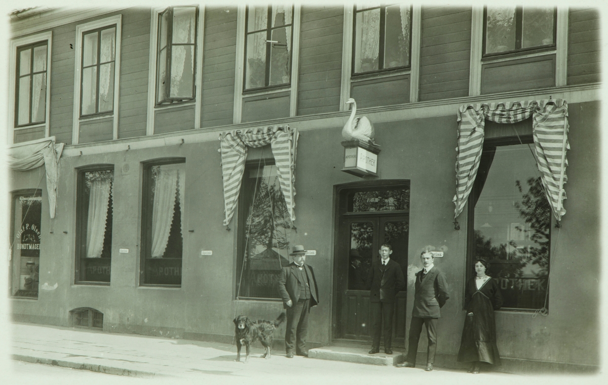 Postkort, Hamar, Strandgata 23, eksteriør Svaneapoteket, til venstre: apoteker Karl Rafael Nikolai Eidem og 3 ansatte, I 1918 flyttet apoteket til Godagergården ved Østre Torg, samme året begynte man å sette opp Festiviteten her. Til venstre holdt Bundtmaker Olaf P. Olsen til,