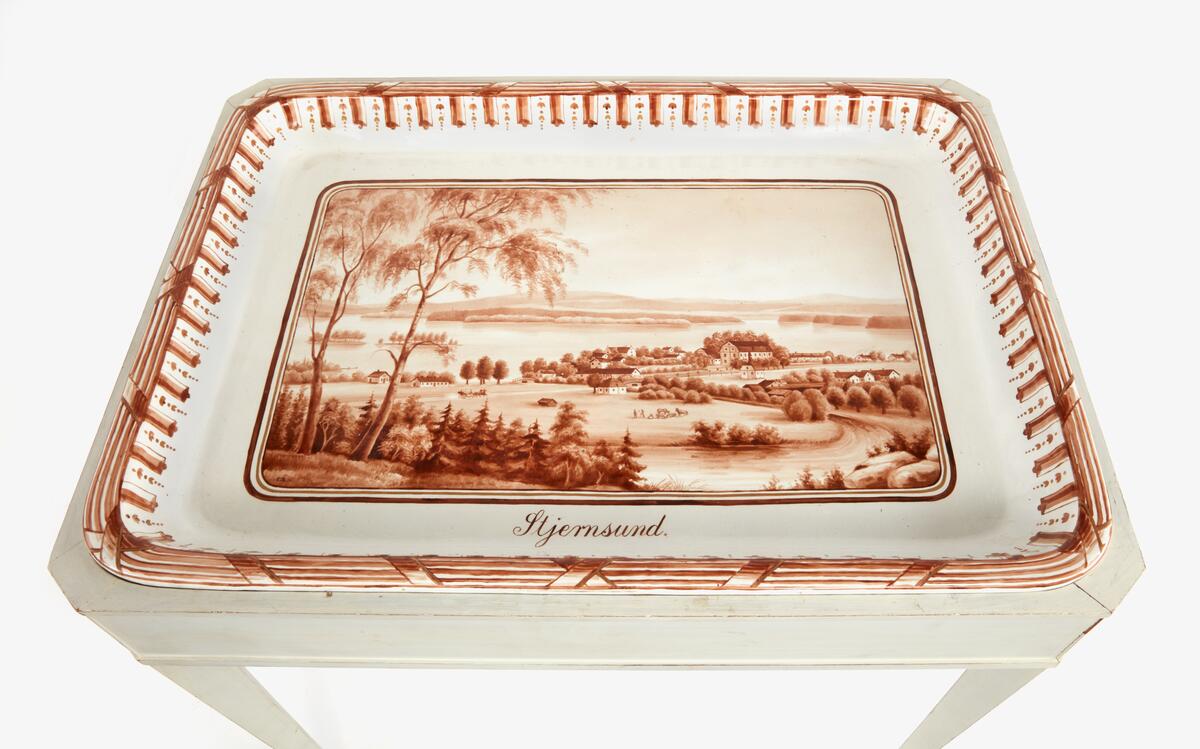 Brickbord - Rörstrand. Lös bricka i porslin med dekor i sepia och guld föreställande Stjernsunds bruk, Dalarna.
Förlaga: Grafiskt blad av Thora Thersner, mitten av 1800-talet. Monogramsignerad och daterad -08 samt märkt Rörstrand. Vitmålat originalunderede i sengustaviansk stil. Möjligen dekorerat av G. J. Asplund, verksam vid Rörstrand 1890-1920.