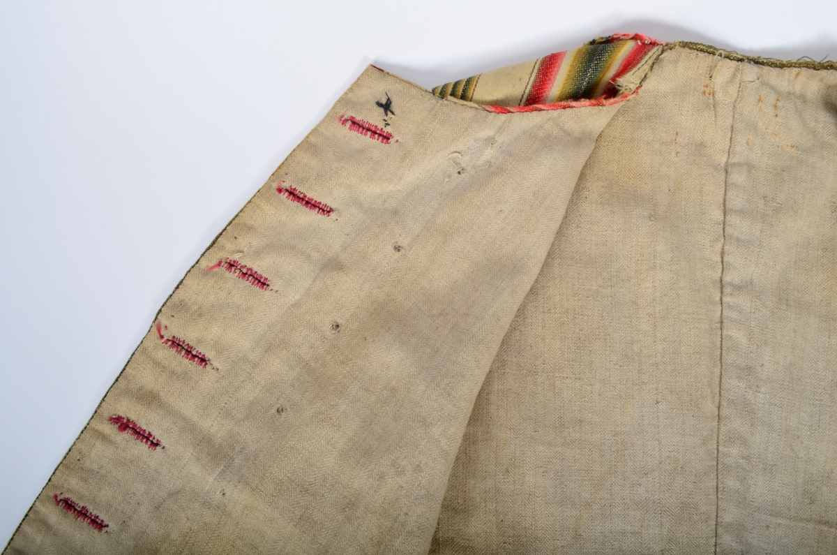 Materiale: 5-skafta satengvoven stripa ullty medrenningseffekt. Innslag av gulkvit ull. Renning i gulkvitt, grønt,
gulgrønt, raudt, lysraudt, lilla og lysbrunt. Stripene er laga slik at fargene går over i hverandre. Knappholstråd i blåraudt
ullgarn. Fôr i spisskypertvove linty. Snitt: Sein rokokko med opne flaster i rygg- ig sidesaum. Dobbeltspent. Knappane
manglar, merke etter dei. Utskrådd framkant nede, svakt bua oppe mot halsringinga. Svakt skrådde ryggstykker med saum
midt i ryggen. Bakovertrekte sidesaumar, skøytt ved sidesaumane. Svakt bakovertrekte skuldersaum. Bua
lommeklaffar på framstykka, men inga lomme. Smal rolu rlangs halsringinga. Regels i ubleika lintråd over flaskene (splitten).
Ekstratekst. Samansying: för og ty er sydde saman kvar for seg. Tyet med attersting, sømromet er så bretta til eine sida. Ryggog
skuldresaumen er så stikken ned med prikkesting frå retta. Fôret er sydd til med kantststing som synes langs framkanten
som små prikkesting.
