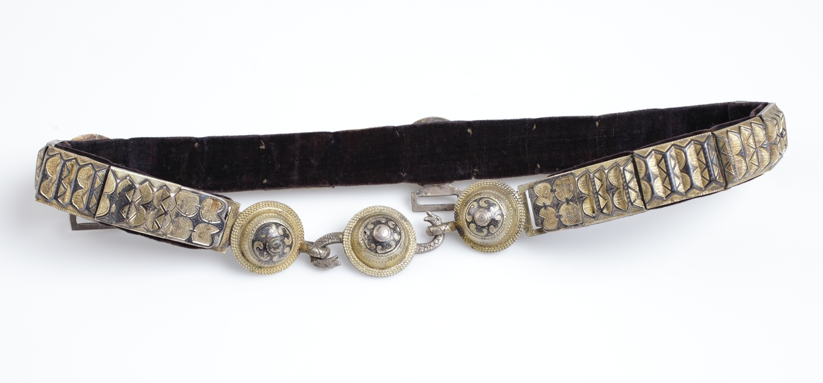 Skärp av metallplåt i guld och svart, framtill tre runda knappar, den mellersta med s-formigt vridna ormhalsar (tjänstgör som lås) de övriga två med öglor, foder av svart sammet. 

Inskrivet i huvudkatalog 1933.