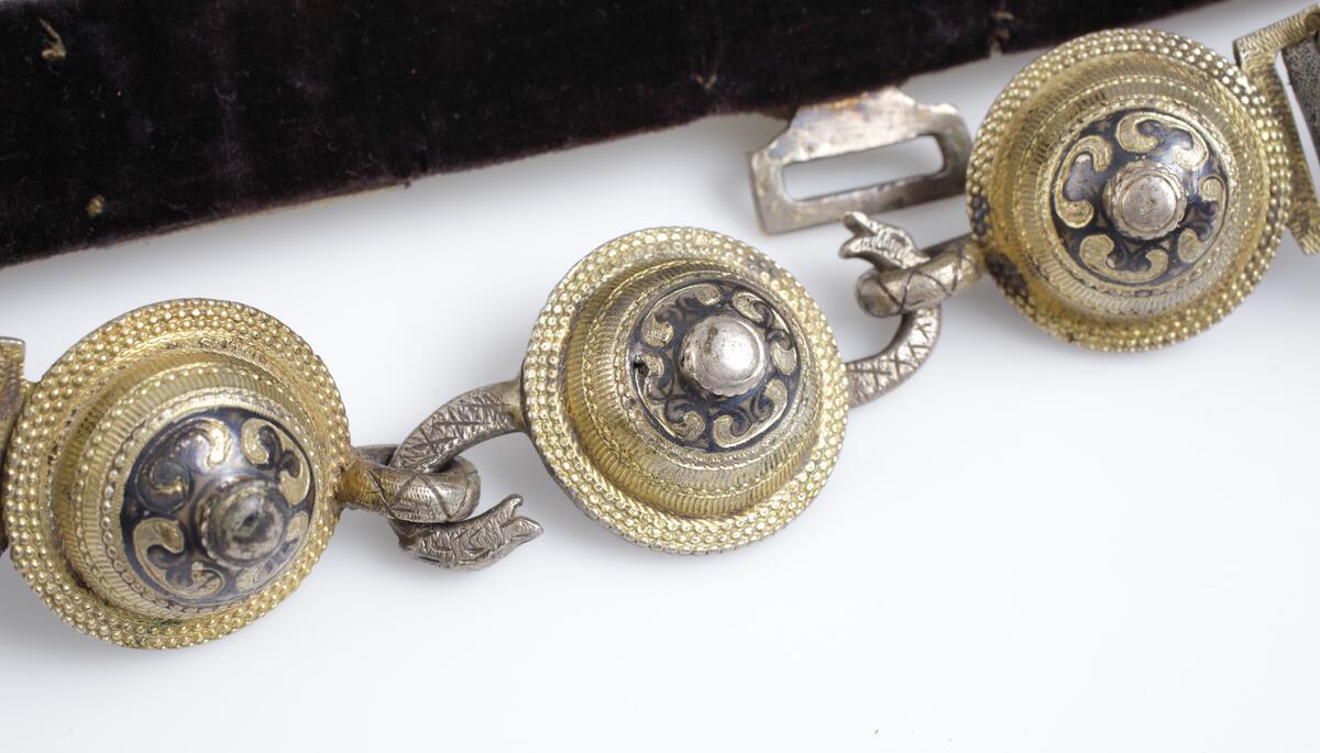 Skärp av metallplåt i guld och svart, framtill tre runda knappar, den mellersta med s-formigt vridna ormhalsar (tjänstgör som lås) de övriga två med öglor, foder av svart sammet. 

Inskrivet i huvudkatalog 1933.