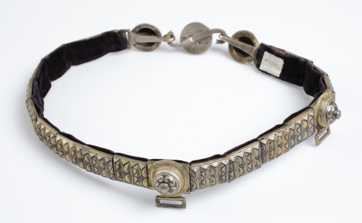 Skärp av metallplåt i guld och svart, framtill tre runda knappar, den mellersta med s-formigt vridna ormhalsar (tjänstgör som lås) de övriga två med öglor, foder av svart sammet. 

Inskrivet i huvudkatalog 1933.