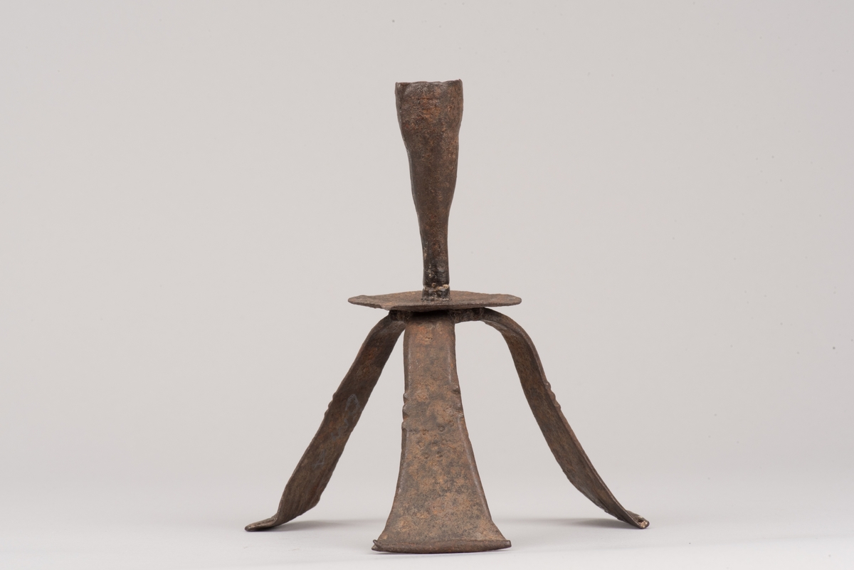 Ljusstake av järnplåt.
Ljusstake med tre fötter som har en uppvikt kant nederst och är avsmalnande uppåt. Fötterna är fästade vid en rund platta där även den smala ljuspipan sitter.
Fötterna är dekorerade med grunt inslagna cirklar som bildar en triangel med spetsarna nedåt. Kanterna är avfasade och ovanför trianglarna är kanten även markerad med några skåror.