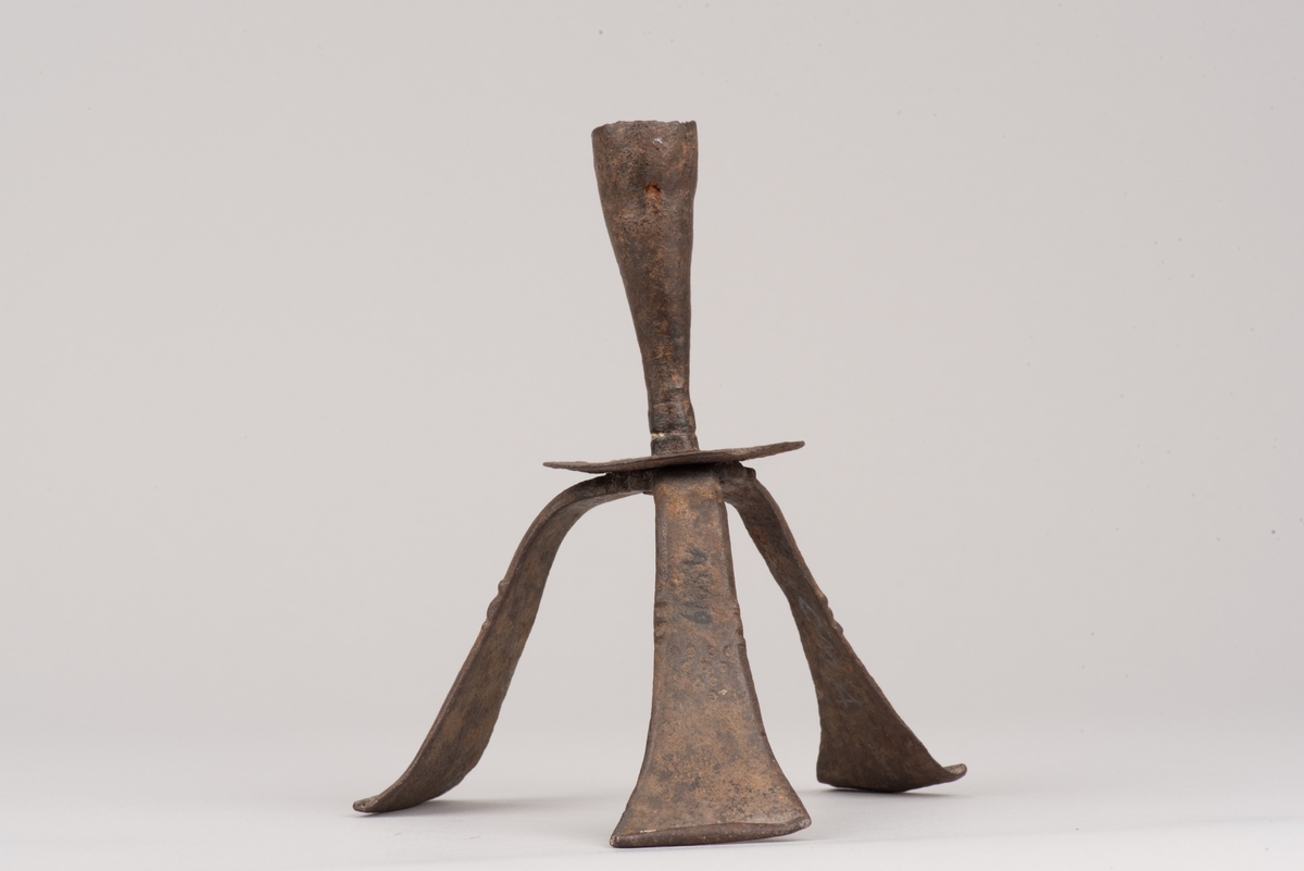 Ljusstake av järnplåt.
Ljusstake med tre fötter som har en uppvikt kant nederst och är avsmalnande uppåt. Fötterna är fästade vid en rund platta där även den smala ljuspipan sitter.
Fötterna är dekorerade med grunt inslagna cirklar som bildar en triangel med spetsarna nedåt. Kanterna är avfasade och ovanför trianglarna är kanten även markerad med några skåror.