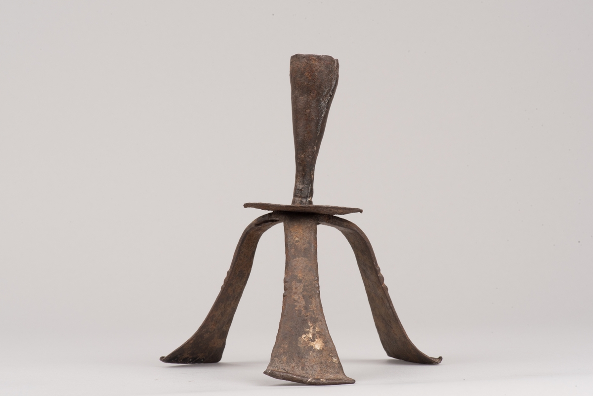 Ljusstake av järnplåt.
Ljusstake med tre fötter som har en uppvikt kant nederst och är avsmalnande uppåt. Fötterna är fästade vid en rund platta där även den smala ljuspipan sitter.
Fötterna är dekorerade med grunt inslagna cirklar som bildar en triangel med spetsarna nedåt. Kanterna är avfasade och ovanför trianglarna är kanten även markerad med några skåror.