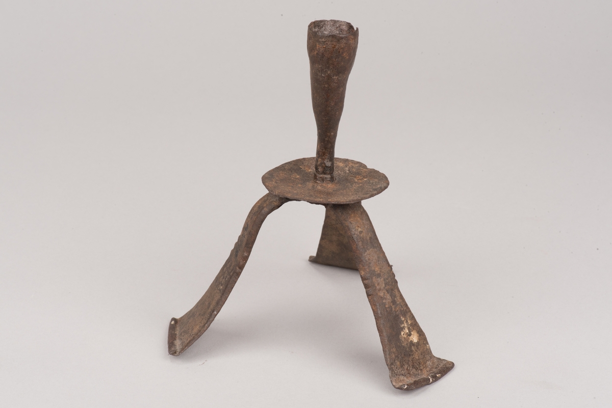 Ljusstake av järnplåt.
Ljusstake med tre fötter som har en uppvikt kant nederst och är avsmalnande uppåt. Fötterna är fästade vid en rund platta där även den smala ljuspipan sitter.
Fötterna är dekorerade med grunt inslagna cirklar som bildar en triangel med spetsarna nedåt. Kanterna är avfasade och ovanför trianglarna är kanten även markerad med några skåror.