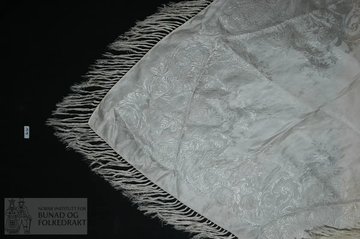 Gulhvit silke med maskinfald i to sider. Påknytte frynser i en rad. Jacquardvevd i blomsterformer langs kantene.  Str.:  74 x 72 cm. Frynser:  11,5 cm.