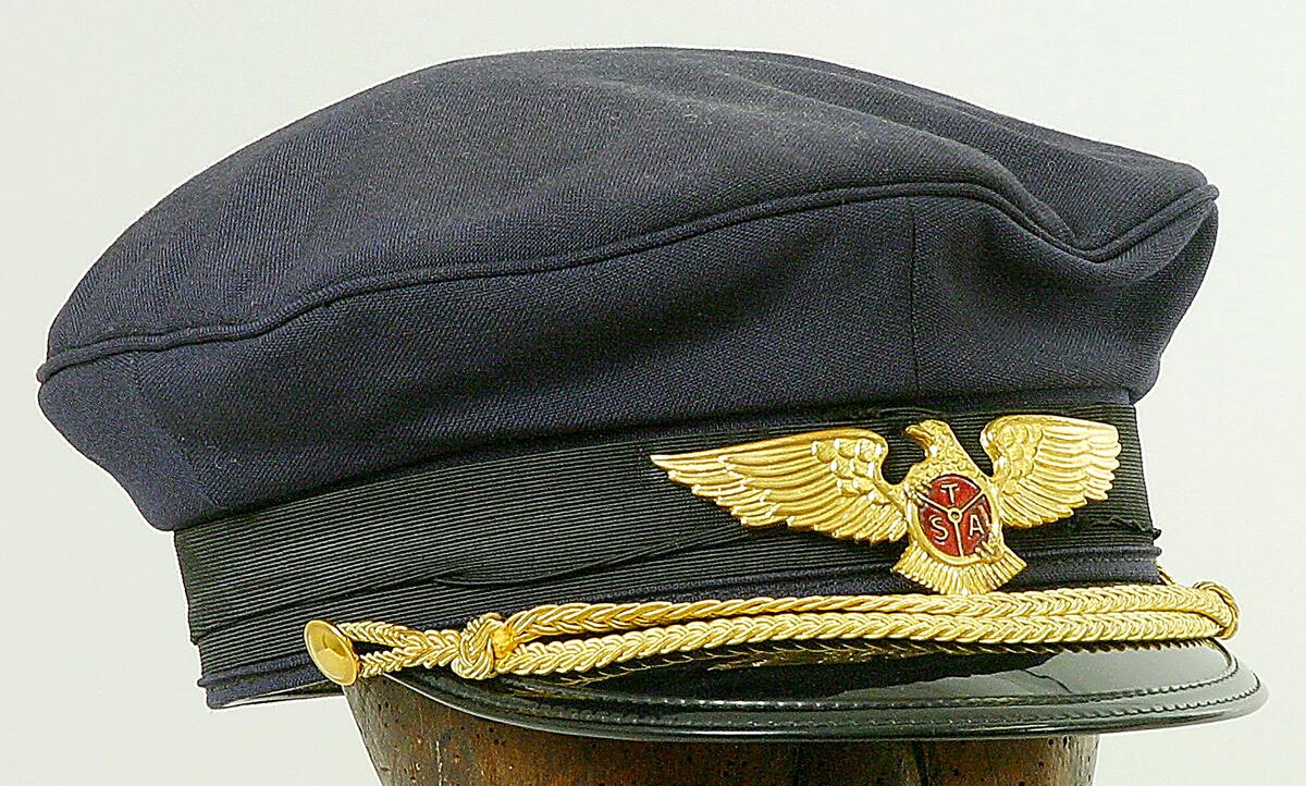 Blå skärmmössa med svart skärm tillhörande trans airs uniform med två flätade guldsnören framtill på skärmen samt en förgylld örnbrosch med bokstäverna t/s/a på röd botten har tillhört lars gibson  som han använde i tjänsten som pilot hos trans air under perioden 1951-1981
nyreg data augusti 2006 lj