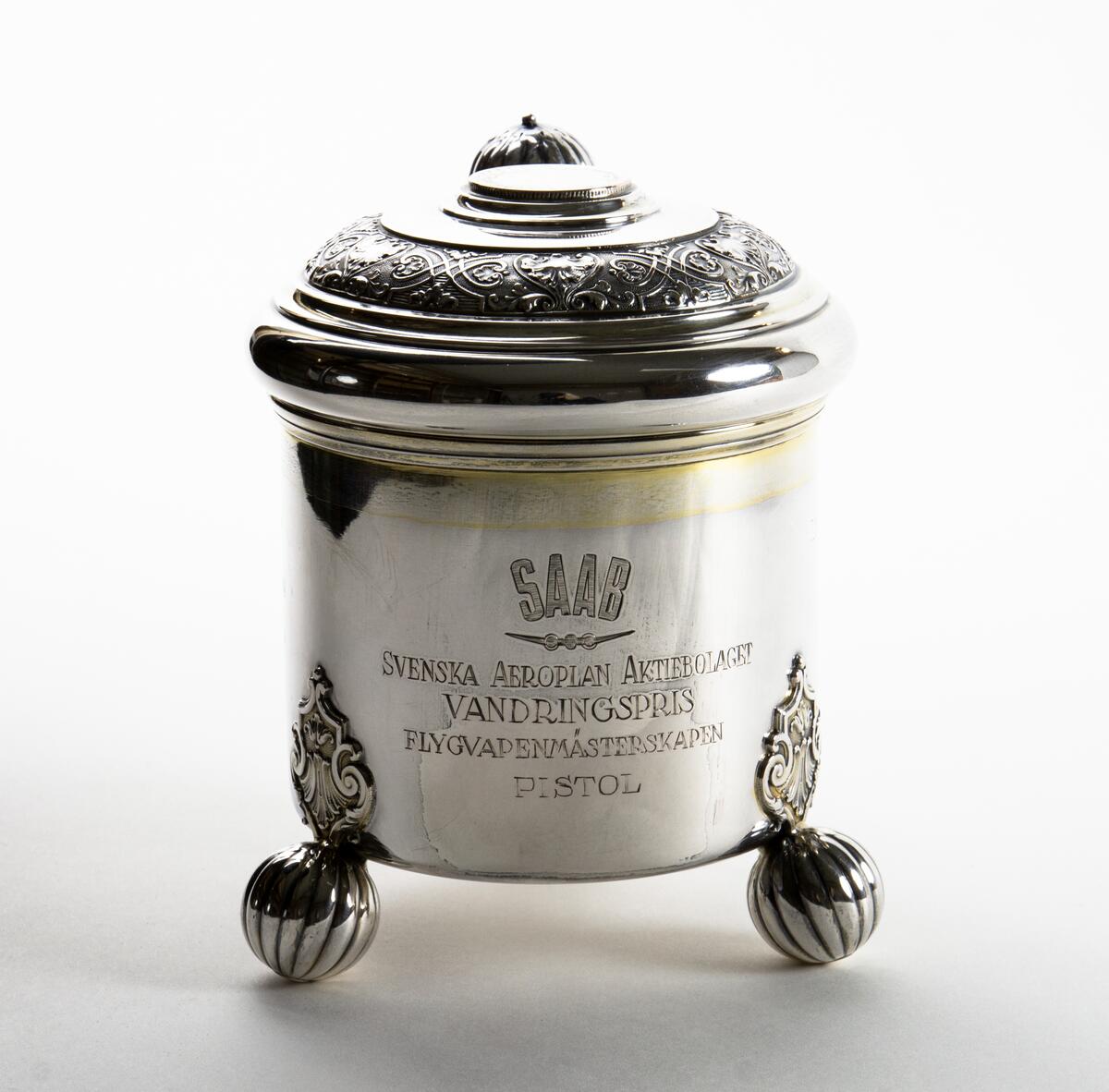 Kanna i silver Prispokal; dryckeskanna i silver Kannan står på tre kulfötter och över fästena för kulfötternafinns en utsmyckning. Tumgreppet som sitter på locket är kulformat. i lockets mitt finns ett mynt, på myntet står det: GUSTAF V SVERIGES KONUNG MED FOLKET FÖR FOSTERLANDET Runt lockets kant finns en utsmyckning. På kannan står det: SAAB SVENSKA AEROPLAN-AKTIEBOLAGET VANDRINGSPRIS FLYGVAPENMÄSTERSKAPEN PISTOL 1948 KUNGL. JÄMTLANDS FLYGFLOTTILJ 1949 KUNGL. VÄSTM. FLYGFLOTTILJ 1950 KUNGL. VÄSTM. FLYGFLOTTILJ 1951 KUNGL. SÖDERM. FLYGFLOTTILJ 1952 KUNGL. SKARAB. FLYGFLOTTILJ 1953 KUNGL. KRIGSFLYGSKOLAN. 1954 KUNGL. SÖDERTÖRNS FLYGFLOTTILJ 1955 KUNGL. KALMAR FLYGFLOTTILJ 1956 KUNGL. VÄSTM. FLYGFOLOTTILJ. Kannan är opolerad.