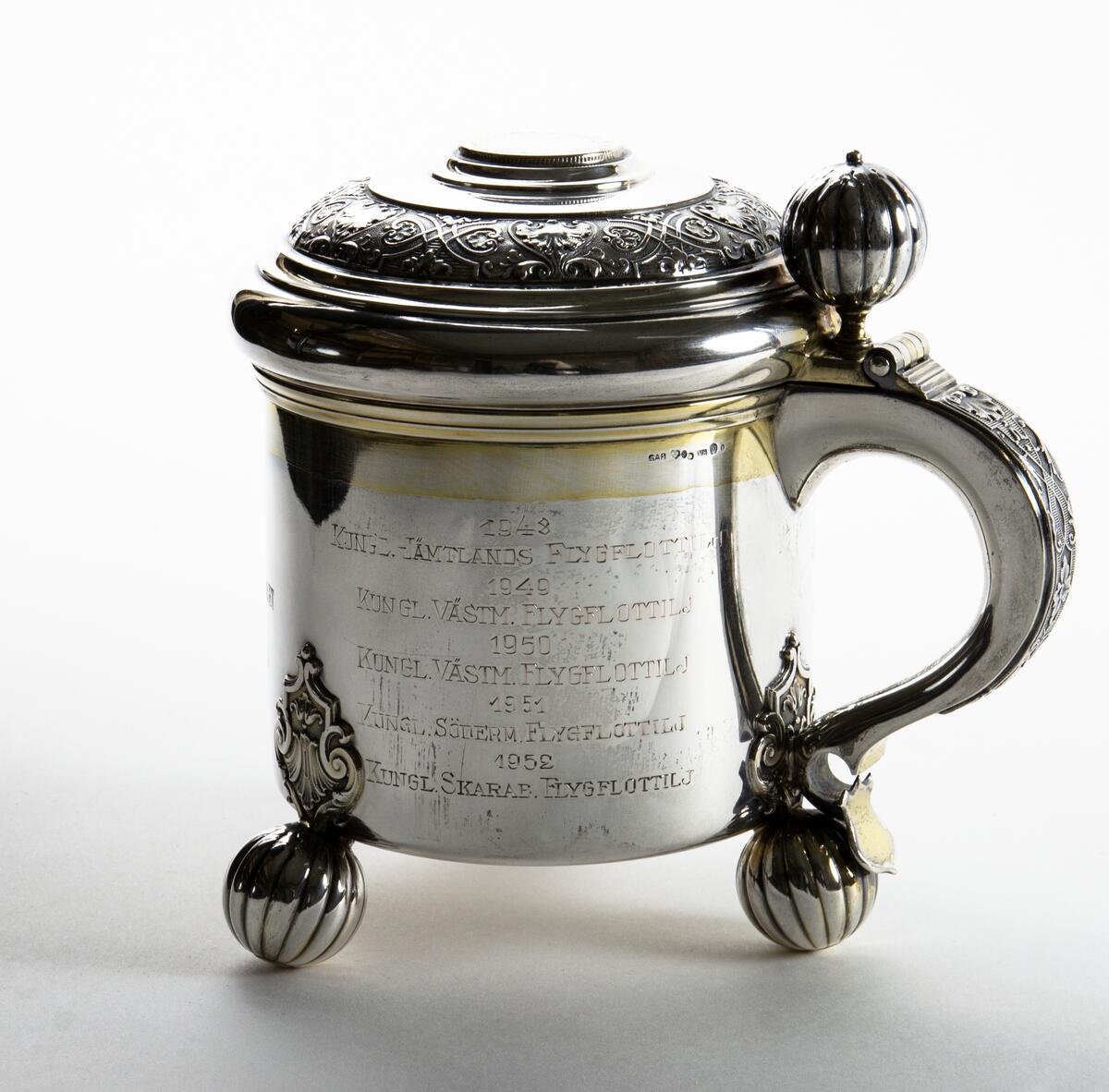Kanna i silver Prispokal; dryckeskanna i silver Kannan står på tre kulfötter och över fästena för kulfötternafinns en utsmyckning. Tumgreppet som sitter på locket är kulformat. i lockets mitt finns ett mynt, på myntet står det: GUSTAF V SVERIGES KONUNG MED FOLKET FÖR FOSTERLANDET Runt lockets kant finns en utsmyckning. På kannan står det: SAAB SVENSKA AEROPLAN-AKTIEBOLAGET VANDRINGSPRIS FLYGVAPENMÄSTERSKAPEN PISTOL 1948 KUNGL. JÄMTLANDS FLYGFLOTTILJ 1949 KUNGL. VÄSTM. FLYGFLOTTILJ 1950 KUNGL. VÄSTM. FLYGFLOTTILJ 1951 KUNGL. SÖDERM. FLYGFLOTTILJ 1952 KUNGL. SKARAB. FLYGFLOTTILJ 1953 KUNGL. KRIGSFLYGSKOLAN. 1954 KUNGL. SÖDERTÖRNS FLYGFLOTTILJ 1955 KUNGL. KALMAR FLYGFLOTTILJ 1956 KUNGL. VÄSTM. FLYGFOLOTTILJ. Kannan är opolerad.