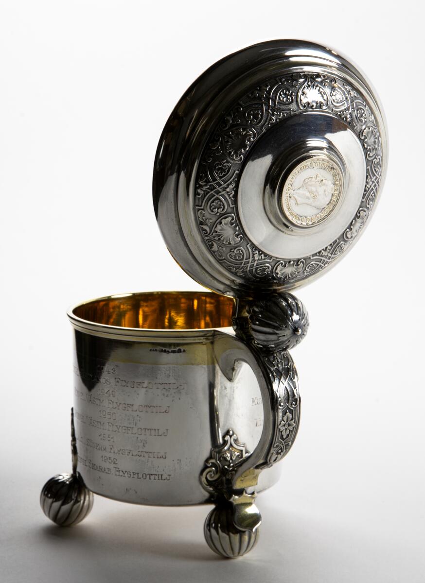 Kanna i silver Prispokal; dryckeskanna i silver Kannan står på tre kulfötter och över fästena för kulfötternafinns en utsmyckning. Tumgreppet som sitter på locket är kulformat. i lockets mitt finns ett mynt, på myntet står det: GUSTAF V SVERIGES KONUNG MED FOLKET FÖR FOSTERLANDET Runt lockets kant finns en utsmyckning. På kannan står det: SAAB SVENSKA AEROPLAN-AKTIEBOLAGET VANDRINGSPRIS FLYGVAPENMÄSTERSKAPEN PISTOL 1948 KUNGL. JÄMTLANDS FLYGFLOTTILJ 1949 KUNGL. VÄSTM. FLYGFLOTTILJ 1950 KUNGL. VÄSTM. FLYGFLOTTILJ 1951 KUNGL. SÖDERM. FLYGFLOTTILJ 1952 KUNGL. SKARAB. FLYGFLOTTILJ 1953 KUNGL. KRIGSFLYGSKOLAN. 1954 KUNGL. SÖDERTÖRNS FLYGFLOTTILJ 1955 KUNGL. KALMAR FLYGFLOTTILJ 1956 KUNGL. VÄSTM. FLYGFOLOTTILJ. Kannan är opolerad.
