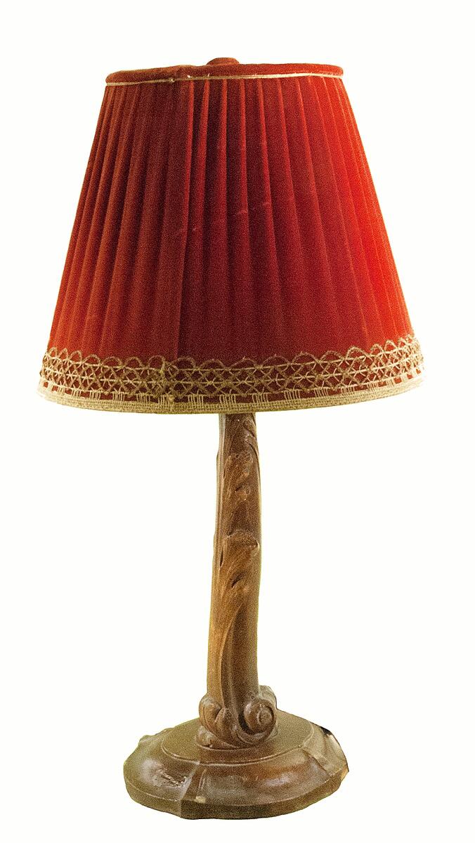 Lampskärm. Till bordslampa. Röd sammet omgärdad av spets i över- och nederkant. Oktagonal, uppåt avsmalnande skärm med plan, veckad topp med centrerad spetsblomma. Stomme och lampfäste av smal vitmålad metall.
Nyreg 20130215 JE