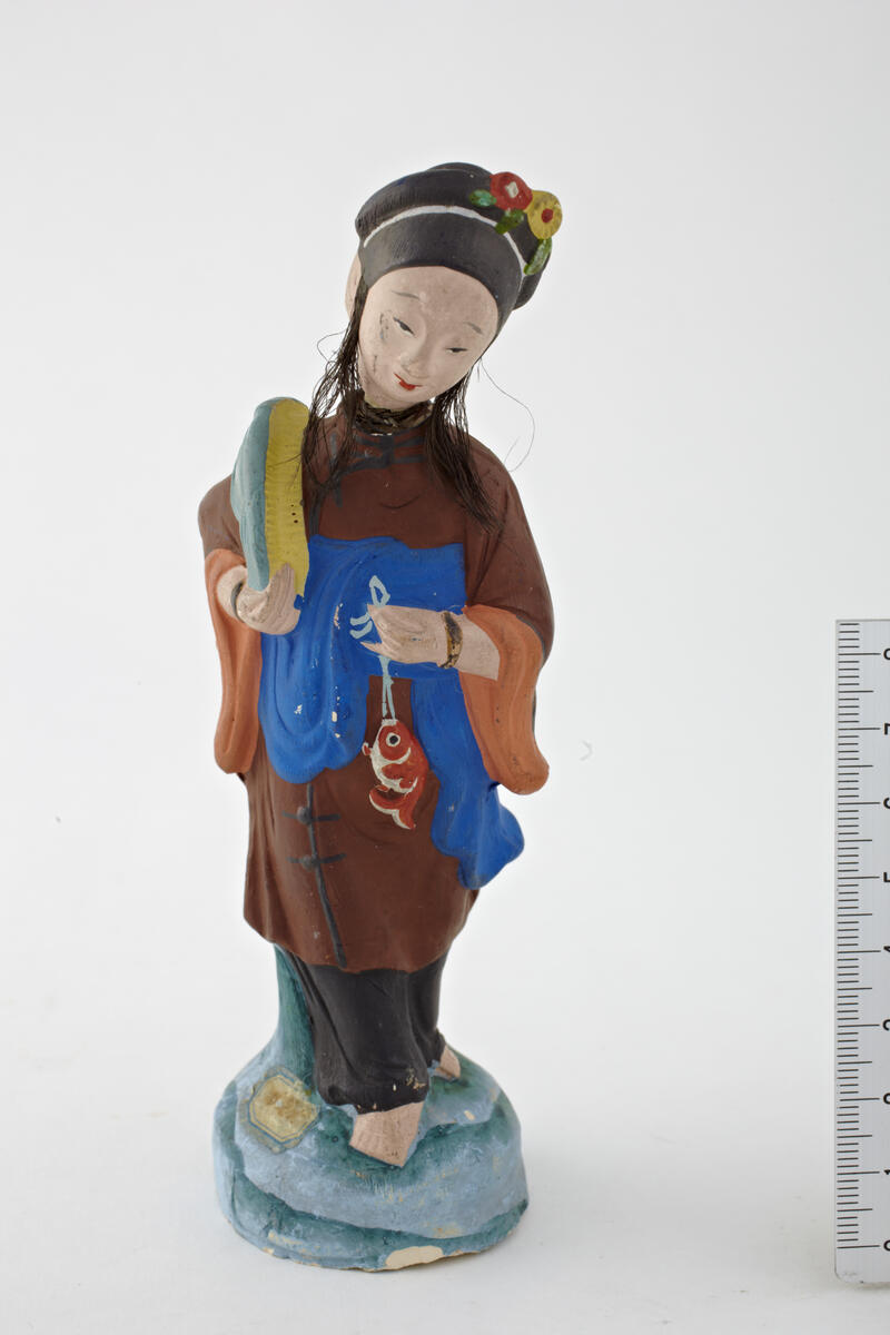 Light grey clay, a lady with real silk hair holding a fish.
Kvindelig figur; med brun kufte; holder i venstre haand, en rød fisk, som hænger i en snor, smlgn. 778