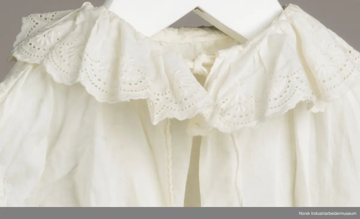 Hvit skjorte med snor i halsen. I kanten av kragen og i enden av ermene er det engelsk blonder.