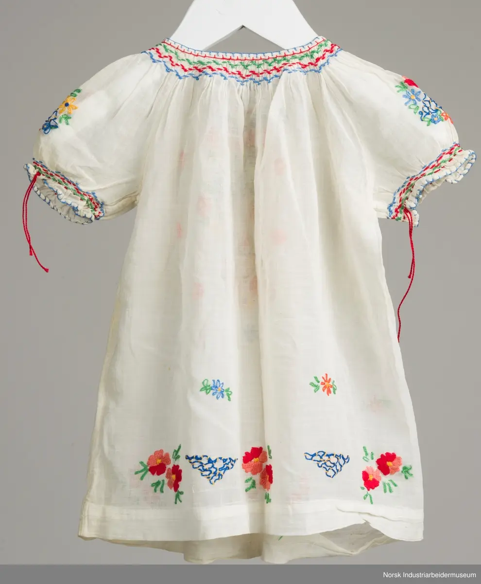 Hvit barnekjole som er brodert med blomstermotiv. Rundt halsen og ermene er det brodert vaffelsøm i grønt, rødt og blått.