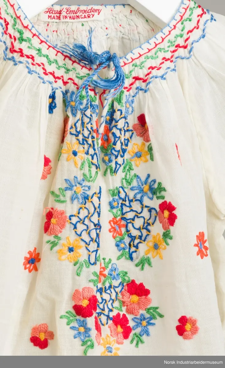 Hvit barnekjole som er brodert med blomstermotiv. Rundt halsen og ermene er det brodert vaffelsøm i grønt, rødt og blått.
