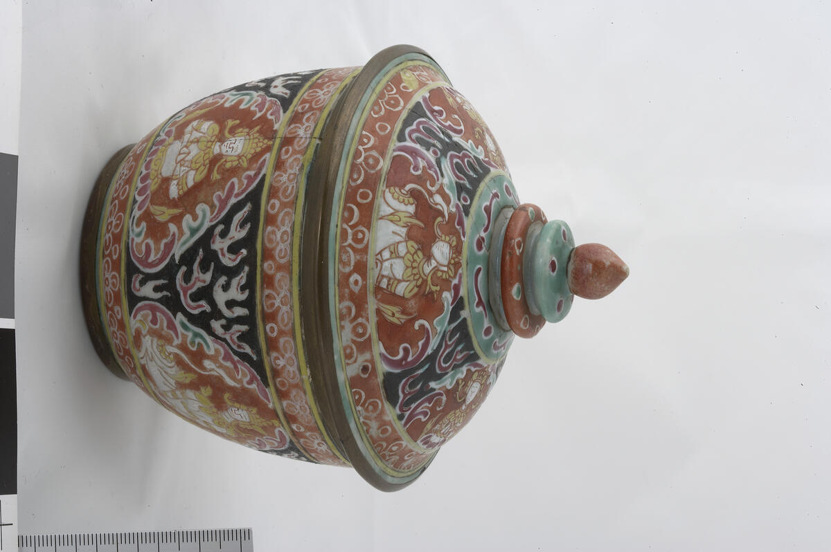 Bencharong. Jar with cover. The deity Theppanom in a triangular medallion is alternating with Norasingh (man-lion) with a halo round his head. Black background with the Buddhist flame, kranok, in pink and white. Red decorated band around the base, rim and cover rim. Green decorated band round the tiered knob, red and green tiers topped by the Buddhist jewel or lotus bud. Red footring. Splayed cover with horizontal rim. Bronze fittings on rim on bowl and cover, and the footring. Jar diameter 10.5 cm, height 7.5 cm. Late 18th to early 19th century.   