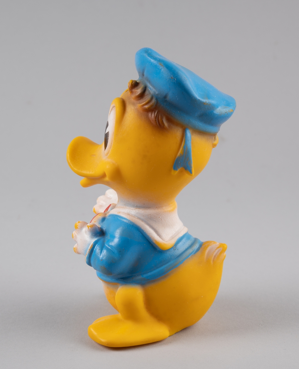 Leketøy av mykplast i form av en Donald Duck. Den er gul med blå jakke med rød sløyfe, den har blå hatt og hvite hansker. Øynene er store og ovale, hvite og svarte.