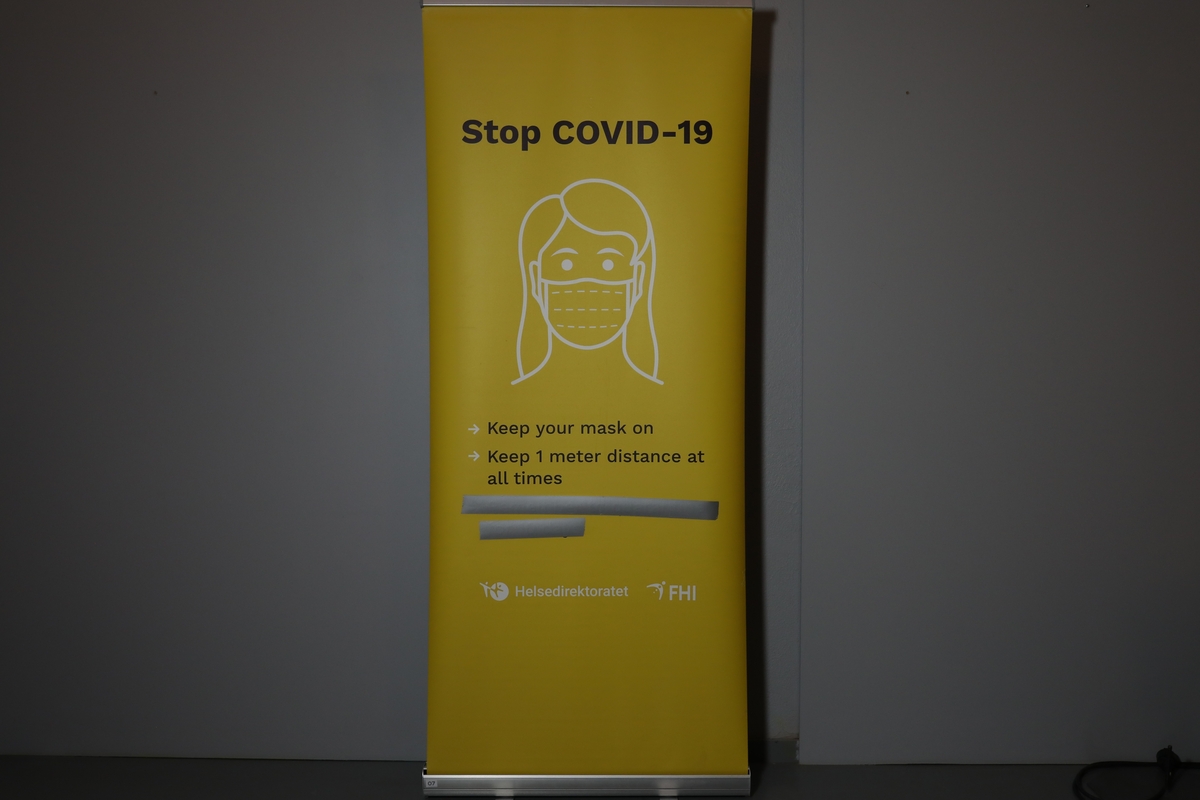 Rollup med med etui, har en stang bak til å holde den oppe. Den har informasjon på engelsk om å bruke maske og holde en meter avstand for å stoppe covid-19. Enkelt bilde av kvinnehodet med ansiktsmaske i mellom tittelen og informasjonen. I bunnen logo til Helsedirektoratet og FHI. Det er teipet over noe tekst i mellom informasjonen og logoene.