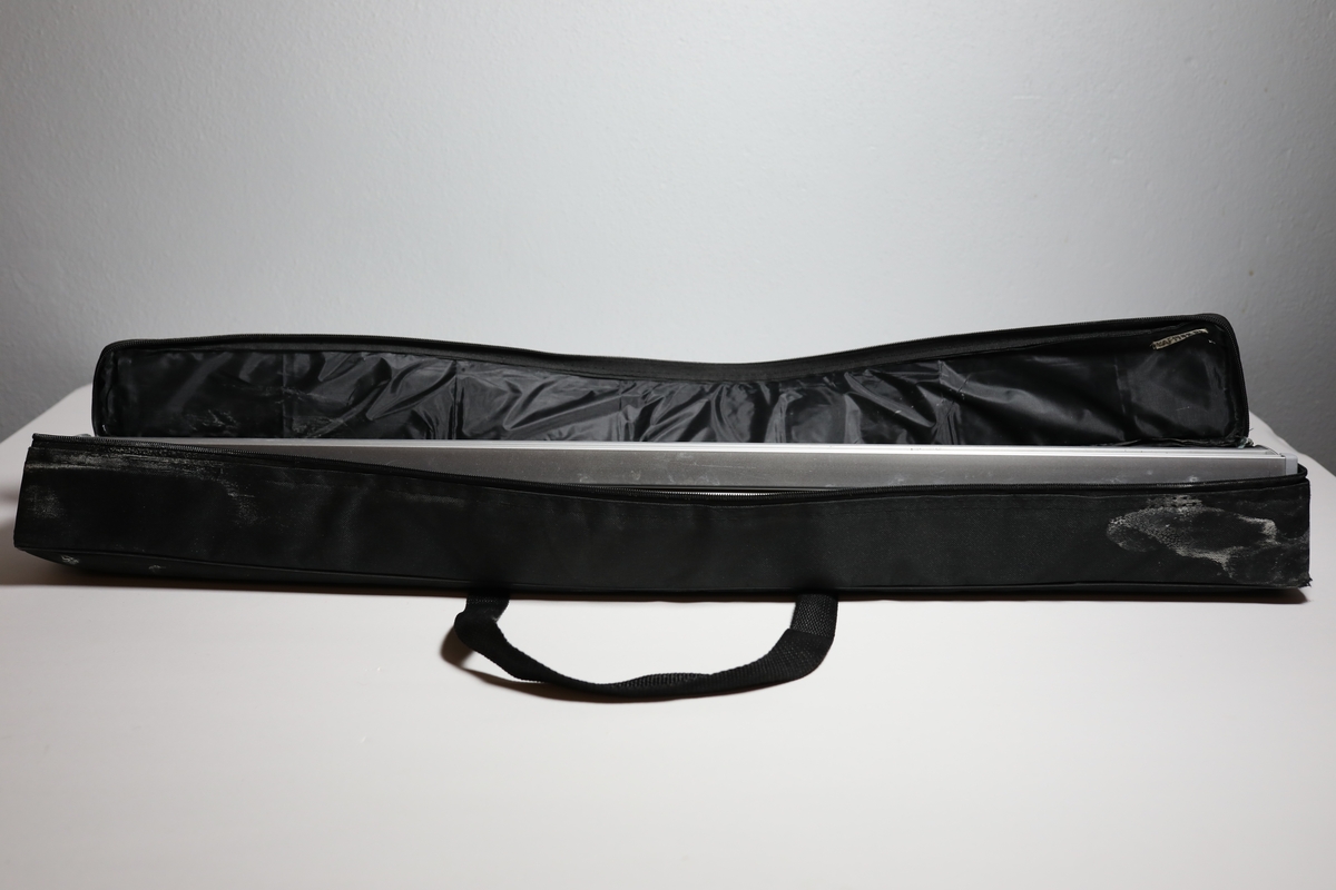 Rollup med med etui, har en stang bak til å holde den oppe. Den har informasjon på engelsk om å bruke maske og holde en meter avstand for å stoppe covid-19. Enkelt bilde av kvinnehodet med ansiktsmaske i mellom tittelen og informasjonen. I bunnen logo til Helsedirektoratet og FHI. Det er teipet over noe tekst i mellom informasjonen og logoene.