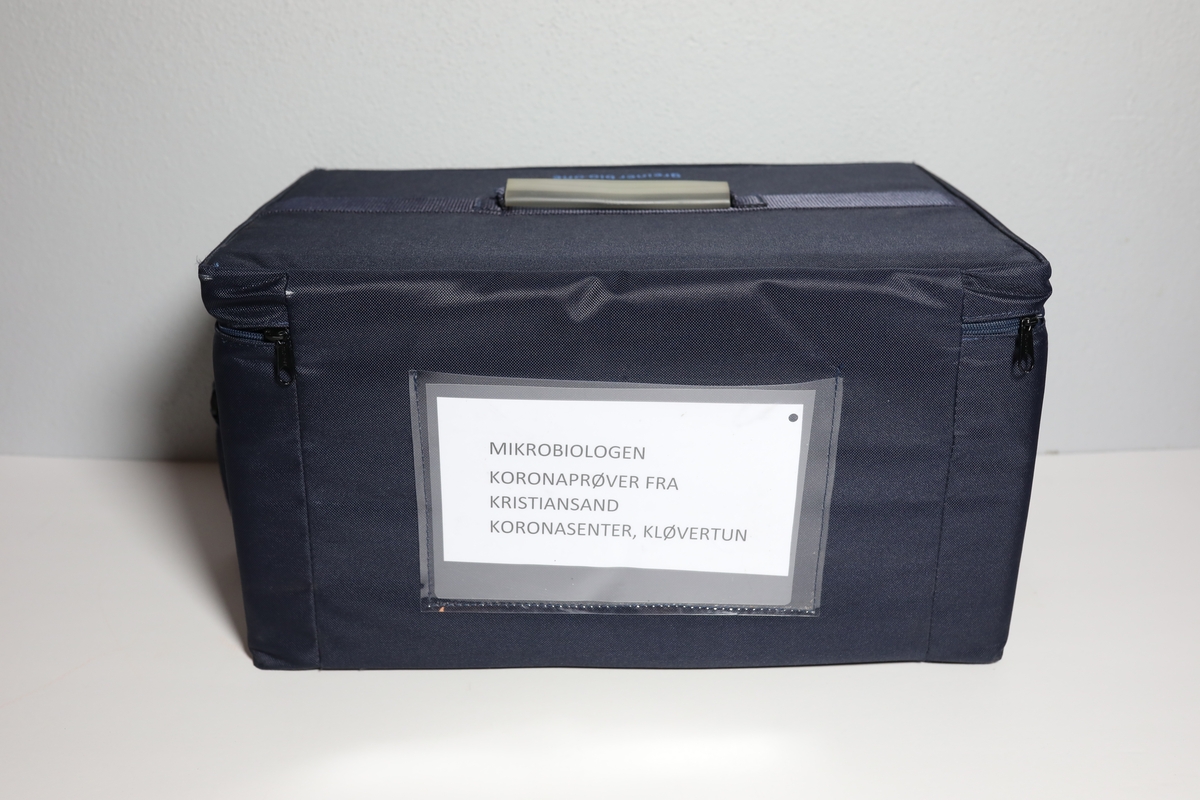 Bag med rom til tre bokser "Vacuette Transport Box" som det ble fraktet Covid-tester i. Glidlås rundt toppen av bagen. Den har håntak i midten av toppen, og på kortside er det feste for strop som man kan bære bagen med. På baksiden er det plastlomme med informasjon. Boksene har gjennomsiktige lokk, og har skumformer i til å plassere opp til 40 testrør i.