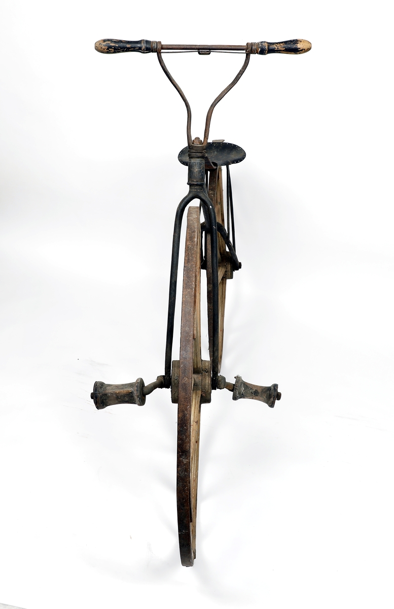 Sykkel av jern med hjul i tre med jernbånd/skoning ytterst. Hjulene har ulik størrelse, størst foran, kan ha vært kjerrehjul en gang, men er laget i smekre materialer. Bemalt i beige med kremhvite linjer, mulig rødt under? Jernrammen er svart, med rester etter dekorasjon i form av linjer. Pedaler på framhjulet (samme system som velte-petter). Trehåndtak. Setet har små huller ytterst som kan ha vært feste for polstring/lær.