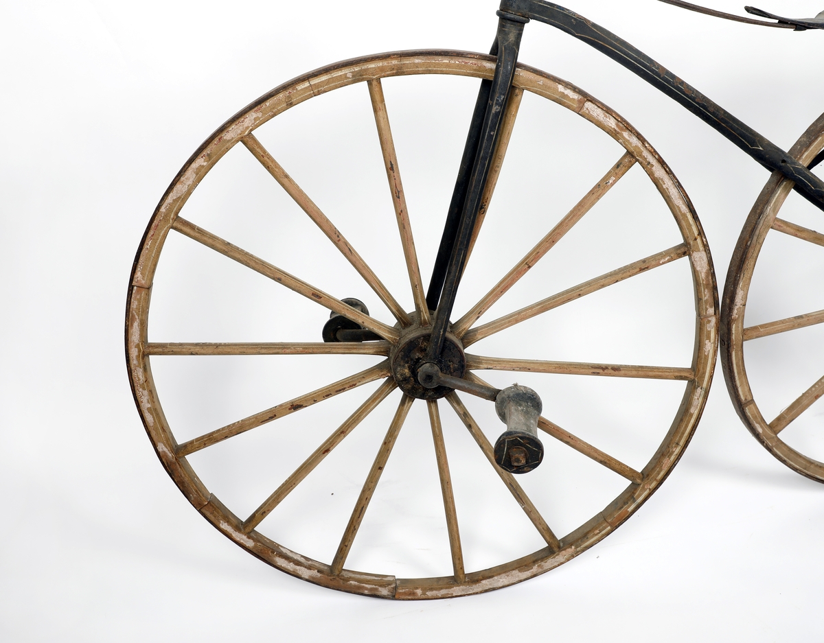 Sykkel av jern med hjul i tre med jernbånd/skoning ytterst. Hjulene har ulik størrelse, størst foran, kan ha vært kjerrehjul en gang, men er laget i smekre materialer. Bemalt i beige med kremhvite linjer, mulig rødt under? Jernrammen er svart, med rester etter dekorasjon i form av linjer. Pedaler på framhjulet (samme system som velte-petter). Trehåndtak. Setet har små huller ytterst som kan ha vært feste for polstring/lær.