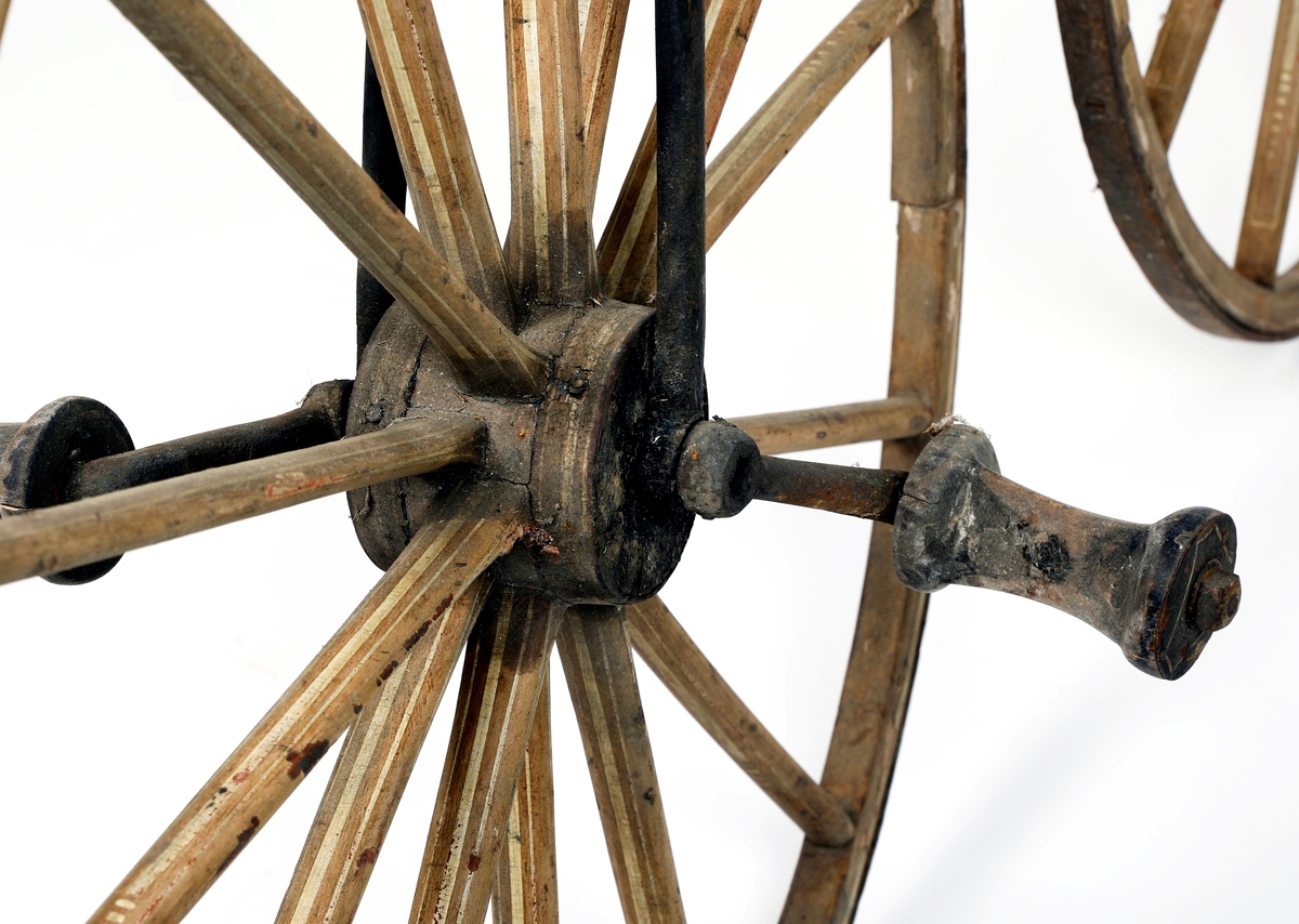 Sykkel av jern med hjul i tre med jernbånd/skoning ytterst. Hjulene har ulik størrelse, størst foran, kan ha vært kjerrehjul en gang, men er laget i smekre materialer. Bemalt i beige med kremhvite linjer, mulig rødt under? Jernrammen er svart, med rester etter dekorasjon i form av linjer. Pedaler på framhjulet (samme system som velte-petter). Trehåndtak. Setet har små huller ytterst som kan ha vært feste for polstring/lær.