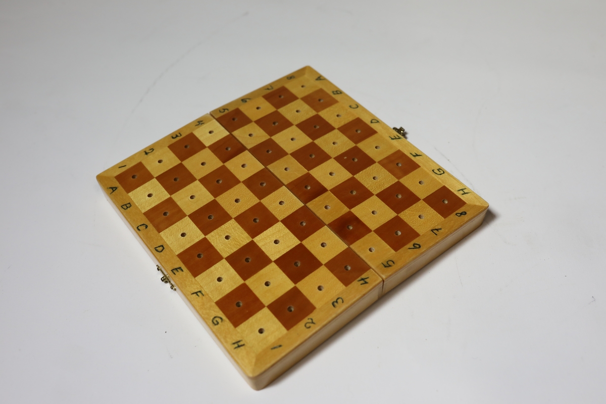 Sjakkbrett i eske med lokk. Rundt kanten er det bokstaver og tall for rutene, og rutene har hull i midten som brikkene kan festes i. Brettet er delt i to og hengslet på midten under. Det er hullrom under med bunn dekket med stoff der brikkene kan oppbevares når brettet er lukket sammen.