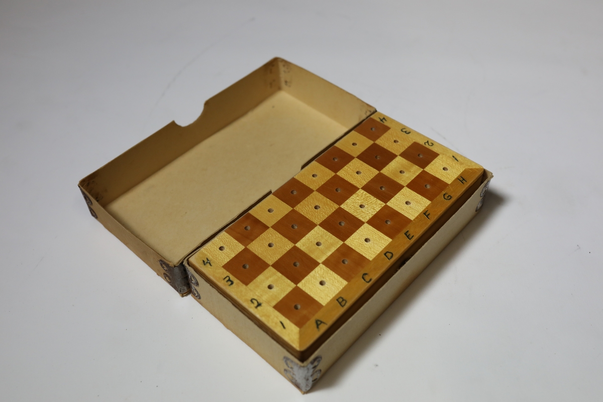 Sjakkbrett i eske med lokk. Rundt kanten er det bokstaver og tall for rutene, og rutene har hull i midten som brikkene kan festes i. Brettet er delt i to og hengslet på midten under. Det er hullrom under med bunn dekket med stoff der brikkene kan oppbevares når brettet er lukket sammen.