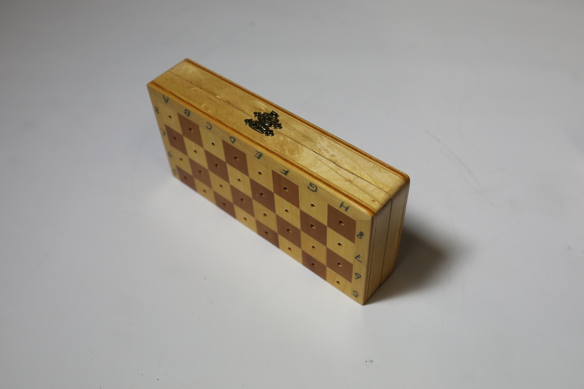 Sjakkbrett i eske med lokk. Rundt kanten er det bokstaver og tall for rutene, og rutene har hull i midten som brikkene kan festes i. Brettet er delt i to og hengslet på midten under. Det er hullrom under med bunn dekket med stoff der brikkene kan oppbevares når brettet er lukket sammen.