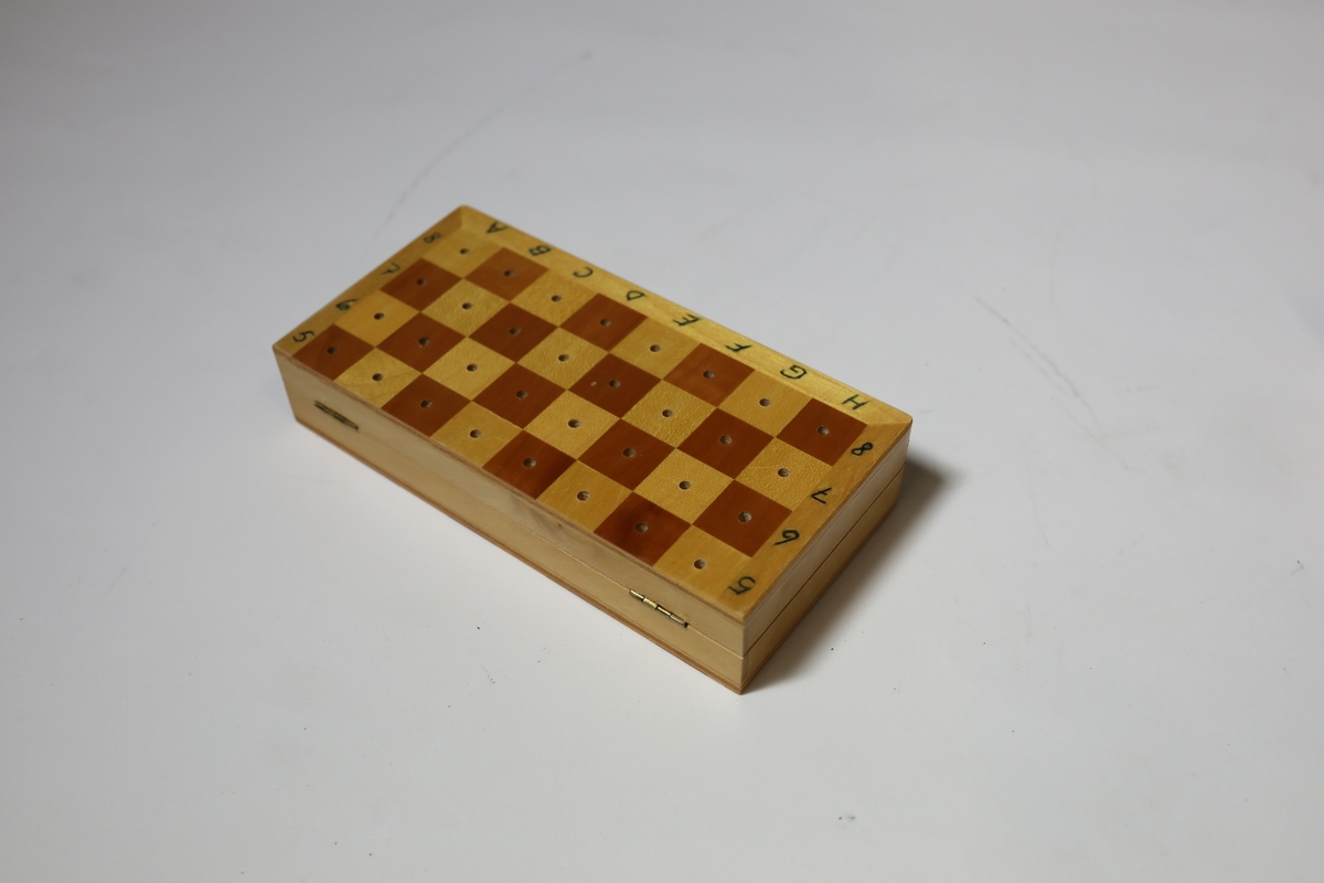 Sjakkbrett i eske med lokk. Rundt kanten er det bokstaver og tall for rutene, og rutene har hull i midten som brikkene kan festes i. Brettet er delt i to og hengslet på midten under. Det er hullrom under med bunn dekket med stoff der brikkene kan oppbevares når brettet er lukket sammen.