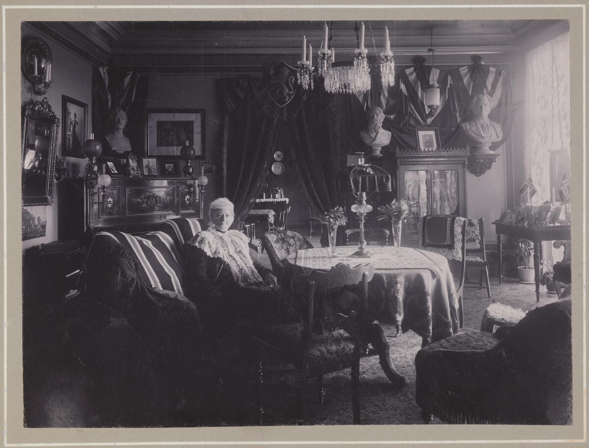 Maren Sars hjemme i salongen på Frogner. Vi finner nasjonale symboler i interiøret: Norske flagg på bordet, bysten av Bjørnstjerne Bjørnson oppe på veggen og portrettene av svigersønnen på pianoet.