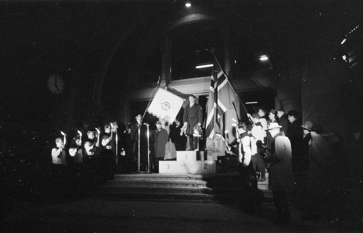 Egil Danielsen kommer hjem til Hamar etter å ha vunnet Olympisk gullmedalje i Spyd i Melbourne 1956. Hamar Jernbanestasjon. Danielsen på toppen av "seierspallen". Pressefotografer, fane Hamar Idrettslag, norsk flagg og fakler. 