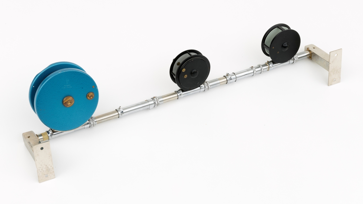 Tre fiskesneller, to fluesneller og en dorge- pilkesnelle, montert på et rør (en stang) med fem snellfester. I og med at det er fem snellefester kan det tyde på at det en gang har vært montert fem sneller på stanga. Snellene er produsert av firmaet Asbjørn Hørgård A/S i Trondheim. I hver ende av røret er det montert fester som gjør det mulig å montere stanga med fiskesneller til vegg. Snellene sitter festet på undersiden av stanga når de brukes, altså med snellefoten opp. 

I salgskatalogen "Råd og redskap fra Hørgård" i 1984 er den blå dorge- pilksnella  beskrevet slik: "Napptun snelle 51/2' * K(EK) Enklere kan en snelle knapt bli. Du kan enten bruke den som pilkesnelle eller du kan dorge med den på sjøen eller etter større innlandsfisk. Snelle har klikk som gir deg et signal når fisken går på. Klikken slår du på ved å skyve på knotten bak. Den store trommelen gjør at det er lett å ta inn fisk. På kanten av spolen har du en bremseflate, som du legger håndflaten på når fiske renner ut. Kan anbefales sammen Frøya. Pris: Napptun m/klikk kr. 205."
Diameter sidedeksel: 14 cm. Bredde målt frfa ytterside av sveivknapper til ytterside av sidedeksel: 8, 2 cm. 

Fluesnellene: Snellene består av spole (trommel), sidedeksler, ramme, sveiv og snellefot. Sveiva er montert direkte på spolen. 
På svart og hvit fluesnelle som er merket Trout Fly sitter sveivknappen løst. Snella merket Trout Fly har disse målene: 
Bredde målt fra ytterside av sveivknapp til ytterside av sidedeksel: 5,6 cm. Bredde spole, innvendig målt: 2,4 cm Diameter sidededeksel: 9,1 cm.
I Hørgård-firmaets bok "Norwegian made fishing tackle" fra 1964 er denne snelletypen beskrevet slik: "Trout Fly 3 1/2" Guttsnelle, passende for flue-, mark- og dorgefiske."

På svart og hvit fluesenlle som er merket Å H Hørgård er disse målene tatt: 
Bredde målt fra ytterside av sveivknapp til ytterside av sidedeksel: 6,4 cm. Bredde spole, innvendig målt: 2,2 cm Diameter sidededeksel: 8,9 cm.
I Hørgård-firmaets bok "Norwegian made fishing tackle" fra 1964 er denne snelletypen beskrevet slik: "Ørret fluesnelle 3 1/2'' ca. 170 gr. Presjonsarbeid med utsøkt materiale. Helstøpt lettmetall. Brennlakkert. Kapasitet: 30 yds. tapered fluesnøre og ca. 50 yds. bakline."



[Se fanen Opplysninger for informasjon om Asbjørn Hørgård og hans firma.]