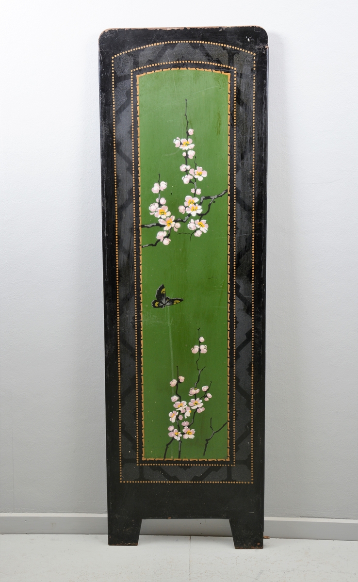 Svart skjermbrett av metall bestående av tre rektangulære paneler som hver står på to føtter. Panelene er dekorert med påmalt dekor i form av grønt vindu med grener med hvite blomster, samt sommerfugl og fugl, innrammet av tre tynne, gullborder.